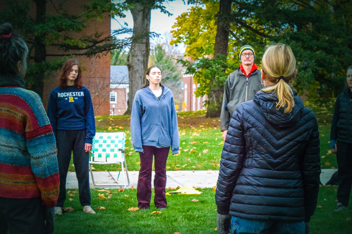 Participants stand in a large outdoor circle with eyes closed, looking upward, engaging in a movement choir activity by the Genesee River.