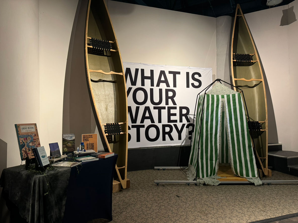Two large wooden canoes stand upright on either side of a green-striped storytelling booth, with a banner in the middle reading “What is your water story?”.