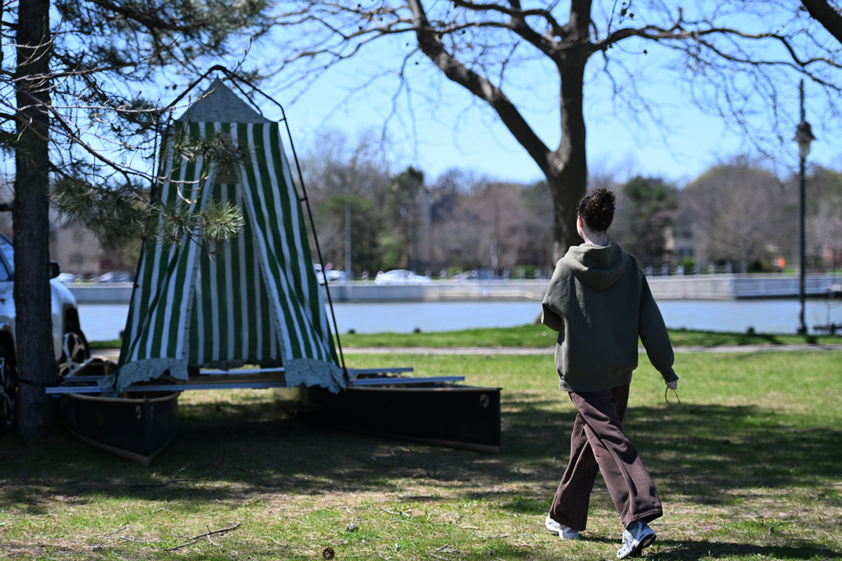 A student walks across the grass toward the green-striped storytelling booth set up near the Genesee River.