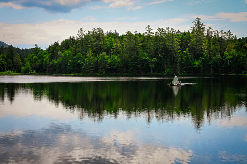 A green-striped storytelling booth set between two canoes is paddled across the calm Raquette River, surrounded by trees at sunset.