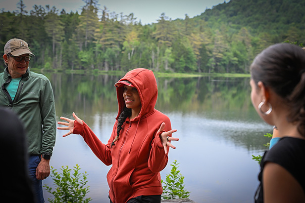 A young woman in a red hoodie gestures expressively while sharing her story with other participants beside the river, with trees and water in the background.